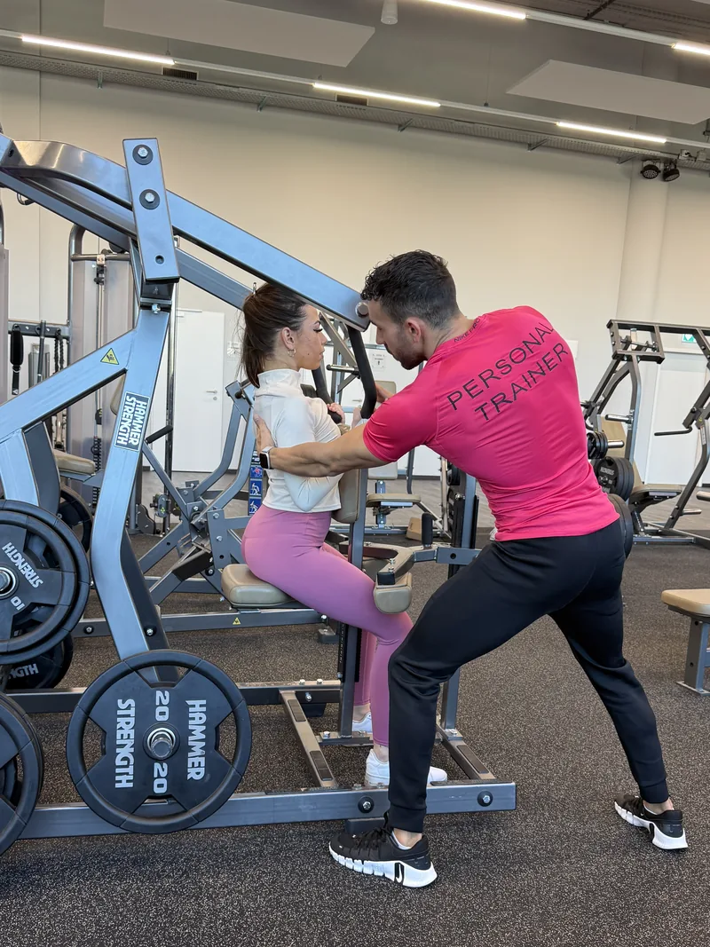 Samy, en t-shirt Personal Trainer, corrige la posture d’une cliente sur une machine de musculation